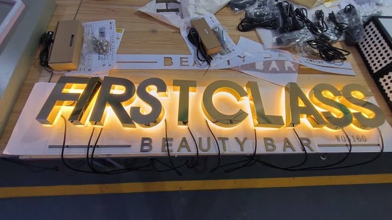 May include: A gold-toned metal sign featuring the words FIRST CLASS in large, illuminated 3D letters with a warm glow, positioned above the text BEAUTY BAR in smaller, non-illuminated letters, with black electrical wires extending from the base.