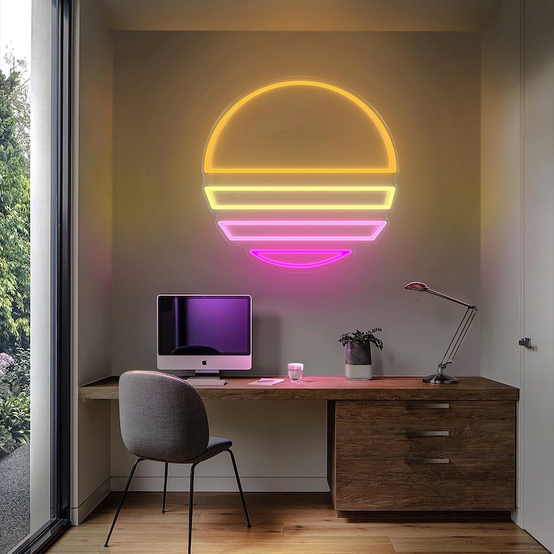 May include: A modern desk with a computer, lamp, and a neon sunset sign on the wall. The desk is made of wood and has a drawer. The computer is a desktop computer with a black screen. The lamp is a desk lamp with a black base and a silver arm. The neon sunset sign is a circular sign with a pink, yellow, and orange gradient.