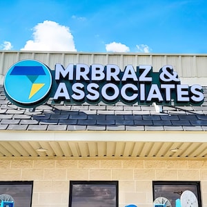 Puede incluir: Letrero de "MRBRAZ & ASSOCIATES" con un logotipo azul y amarillo. El letrero está montado en un edificio con un tejado gris y una fachada de color claro. El cielo es azul con algunas nubes.