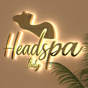 May include: A gold-colored illuminated sign for a spa, featuring the word "Headspa" in elegant script, with "Italy" below. The sign includes a silhouette of a person's head and shoulders. The sign is mounted on a light-colored wall.
