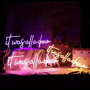 May include: Two neon signs with the text "it was all a dream" in white lettering. The top sign is purple and the bottom sign is pink. The signs are on a table with books and a decorative box.