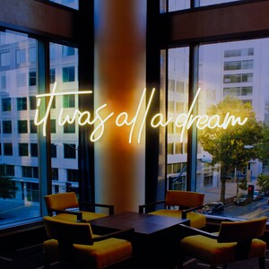 May include: A neon sign with the text "it was all a dream" hanging in a modern office space with large windows. The sign is illuminated with a warm yellow glow. There are four chairs and a table in the room.