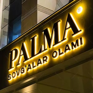 May include: Illuminated sign for "PALMA SOVG'ALAR OLAMI" in a nighttime setting. The sign features bold, black lettering with a bright yellow glow, creating a striking contrast against the dark background. The design includes a registered trademark symbol.