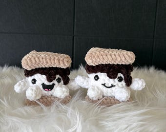 Könnte beinhalten: Zwei handgefertigte S'mores-Spielzeuge aus Häkelarbeit. Jedes hat eine obere Schicht aus beigefarbenen Graham-Crackern, eine dunkelbraune Schokoladenschicht und einen weißen Marshmallow-Körper mit einem lächelnden Gesicht und kleinen Armen. Sie werden auf einer flauschigen weißen Oberfläche präsentiert.