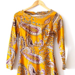 May include: A yellow dress with a paisley print. The dress has long sleeves and a round neckline.