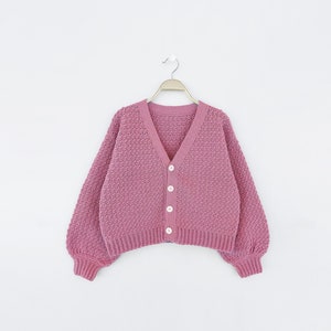May include: A dusty rose-colored cardigan with a V-neck and long, puffy sleeves. The knitted sweater has a textured pattern and is fastened with three white buttons. The garment is hanging on a metal hanger against a white background.