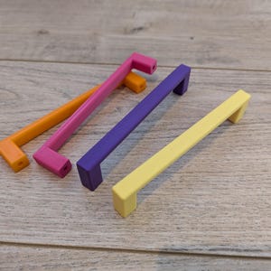 Peut inclure: Quatre poignées de meubles colorées en orange, rose, violet et jaune. Chaque poignée a une barre rectangulaire et une poignée en angle droit. Les poignées sont en matériau solide et sont conçues pour les tiroirs ou les armoires.