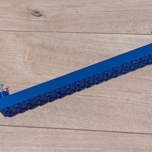 Puede incluir: Un bloque de construcción de plástico azul, largo y recto, con múltiples puntos de conexión. Dos tornillos plateados están unidos a los extremos. El bloque está sobre una superficie de madera marrón claro.