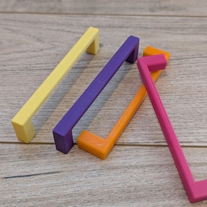 K&ouml;nnte beinhalten: Vier farbenfrohe Schrankgriffe in Gelb, Lila, Orange und Pink. Jeder Griff hat einen rechteckigen Stab und einen rechtwinkligen Griff. Die Griffe sind auf einer hellbraunen Holzoberfl&auml;che angeordnet.