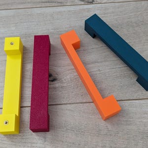 Peut inclure: Quatre poignées de meubles colorées en jaune, magenta, orange et bleu canard. Les poignées sont rectangulaires avec une poignée qui s'étend de chaque extrémité. La poignée jaune a une tête de vis visible. Elles sont disposées sur une surface en bois clair.