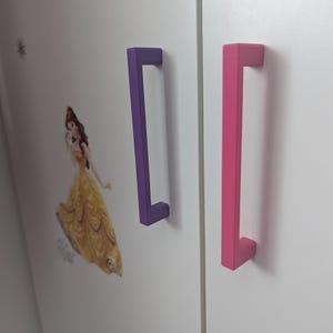 K&ouml;nnte beinhalten: Eine wei&szlig;e Schrankt&uuml;r mit einem lila und einem rosa rechteckigen Griff. Ein gelbes Kleid und ein Prinzessinnen-Aufkleber befinden sich auf der Schrankt&uuml;r.