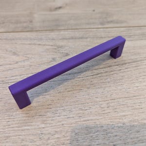 K&ouml;nnte beinhalten: Ein durchgehender violetter Schrankgriff mit einem rechteckigen Stab und zwei rechtwinkligen St&uuml;tzen. Die Hardware ist f&uuml;r eine einfache Installation konzipiert und eignet sich f&uuml;r Schubladen und Schr&auml;nke. Der Griff ist etwa 15 cm lang.