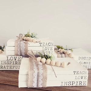 May include: Stack of white books with burlap and lace ribbon, wooden beads, and greenery. The books have the words "LOVE INSPIRE GRATEFUL", "TEACH LOVE INSPIRE", and "DECIDE COMMIT SEED TEACH LOVE INSPIRE" printed on them.