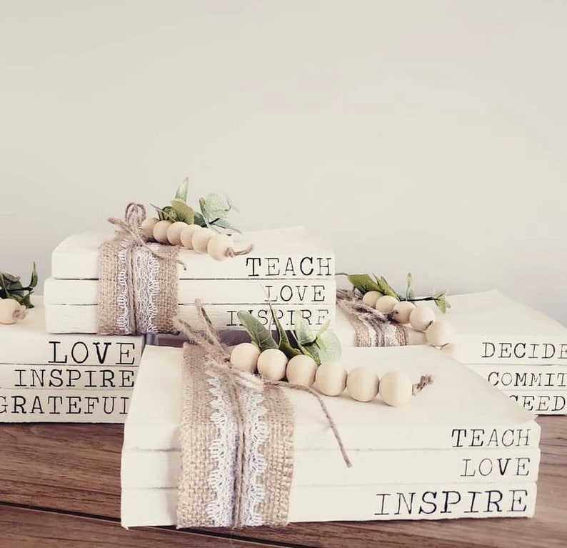 May include: Stack of white books with burlap and lace ribbon, wooden beads, and greenery. The books have the words "LOVE INSPIRE GRATEFUL", "TEACH LOVE INSPIRE", and "DECIDE COMMIT SEED TEACH LOVE INSPIRE" printed on them.