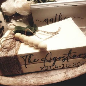 May include: Two white books stacked on top of each other. The top book has the text "The Agostinos" in black script font. The bottom book has the text "EST. 4-10-2021" in black font. The books are decorated with a brown burlap ribbon and a string of wooden beads.