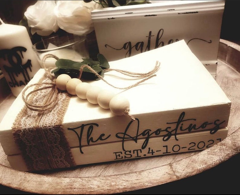 May include: Two white books stacked on top of each other. The top book has the text "The Agostinos" in black script font. The bottom book has the text "EST. 4-10-2021" in black font. The books are decorated with a brown burlap ribbon and a string of wooden beads.