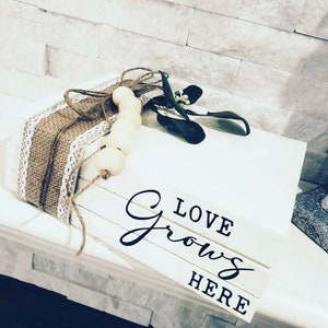 May include: A stack of white wooden blocks with the words "LOVE GROWS HERE" written in black. The blocks are decorated with a brown burlap ribbon and a string of white wooden beads.