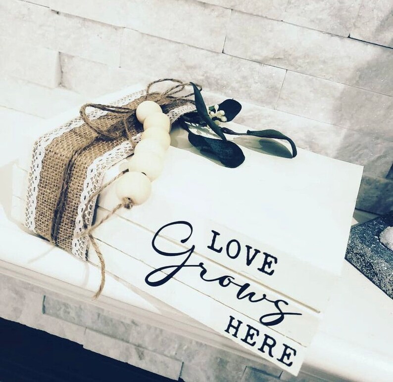 May include: A stack of white wooden blocks with the words "LOVE GROWS HERE" written in black. The blocks are decorated with a brown burlap ribbon and a string of white wooden beads.