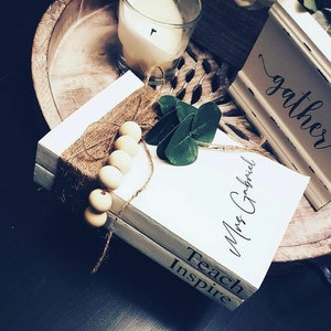 May include: Two white books stacked on top of each other. The top book has the text "Mrs. Gabriel" written on it in black cursive. The bottom book has the text "Teach Inspire" written on it in black. The books are decorated with brown twine and wooden beads.
