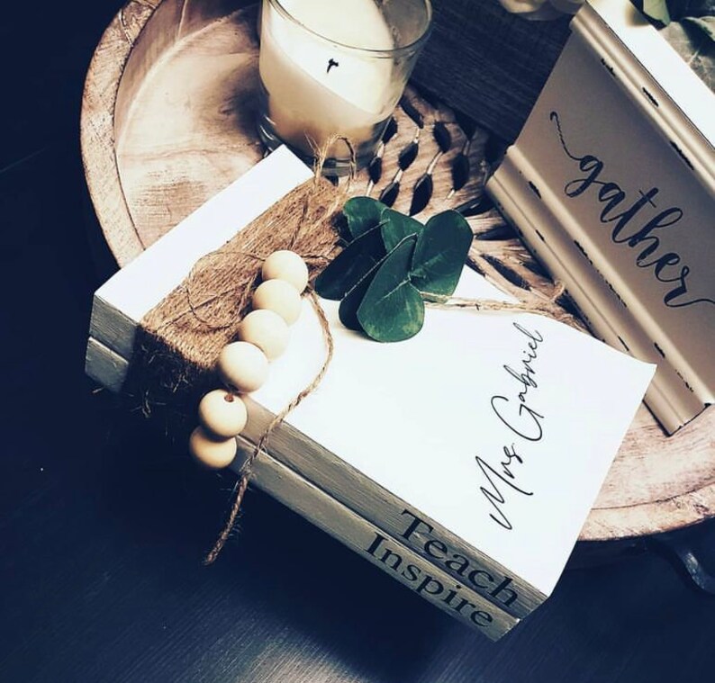 May include: Two white books stacked on top of each other. The top book has the text "Mrs. Gabriel" written on it in black cursive. The bottom book has the text "Teach Inspire" written on it in black. The books are decorated with brown twine and wooden beads.