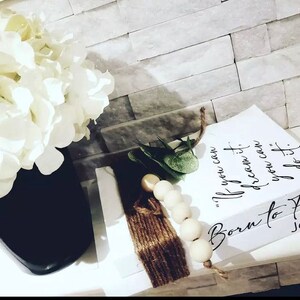 May include: White stacked books with the text "If you can dream it, you can do it." and "Born to Fly" with the name Jane on the bottom book. The books are decorated with a brown tassel and white beads.