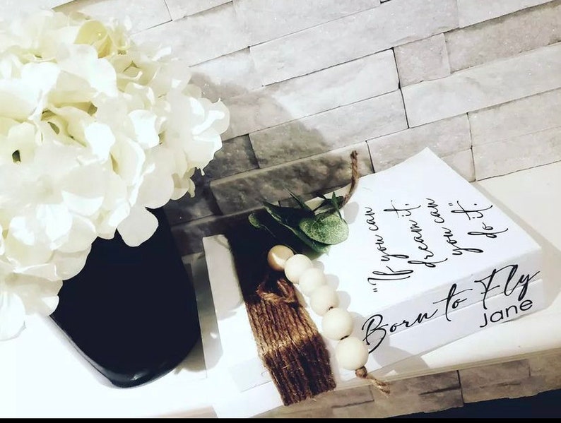 May include: White stacked books with the text "If you can dream it, you can do it." and "Born to Fly" with the name Jane on the bottom book. The books are decorated with a brown tassel and white beads.