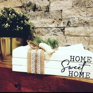 May include: White stacked books with the text "HOME Sweet HOME" in black lettering. The books are decorated with burlap and lace ribbon and a small sprig of greenery with white beads.