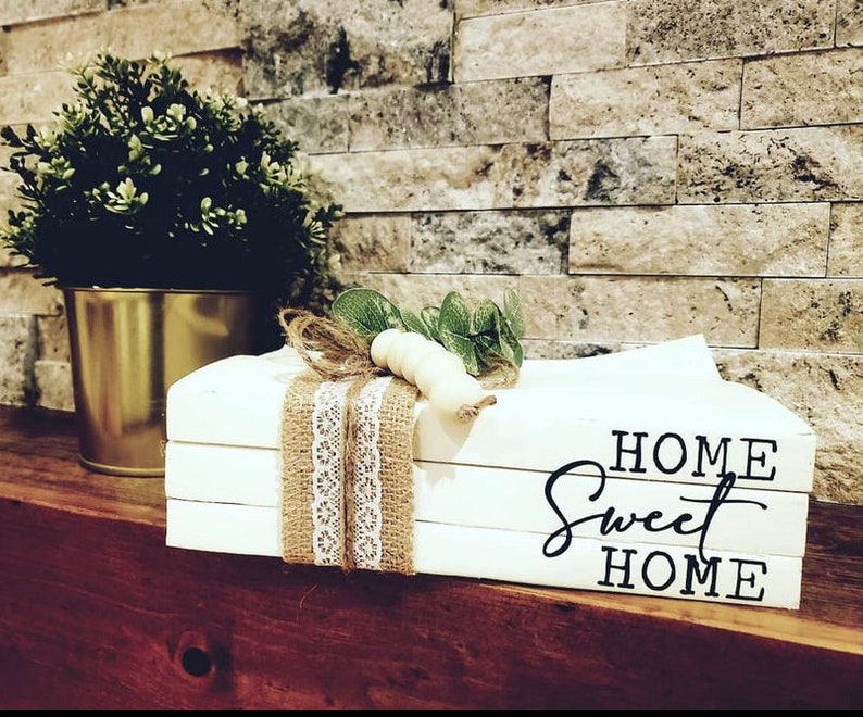 May include: White stacked books with the text "HOME Sweet HOME" in black lettering. The books are decorated with burlap and lace ribbon and a small sprig of greenery with white beads.