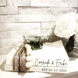 May include: Two white books stacked on top of each other. The top book has the text "Corrado & Erika EST 10-10-2020" in black script font. The books are decorated with a brown burlap ribbon, white lace, and a string of white wooden beads.