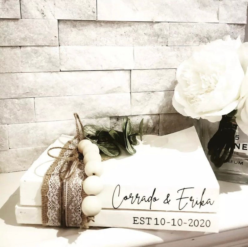 May include: Two white books stacked on top of each other. The top book has the text "Corrado & Erika EST 10-10-2020" in black script font. The books are decorated with a brown burlap ribbon, white lace, and a string of white wooden beads.