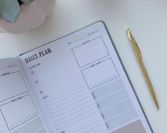 May include: A gray daily planner with a pink and gray interior. The planner has sections for daily tasks, to-do's, self-care, and notes. The planner also has a section for gratitude and fitness & water.