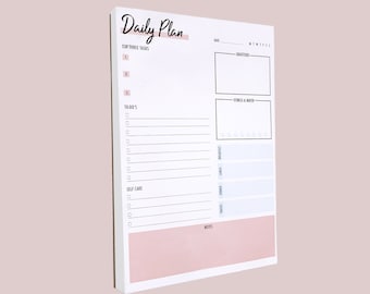 May include: A white daily planner with a pink background. The planner has sections for top three tasks, to-do's, self care, gratitude, fitness & water, notes, and a daily tracker.