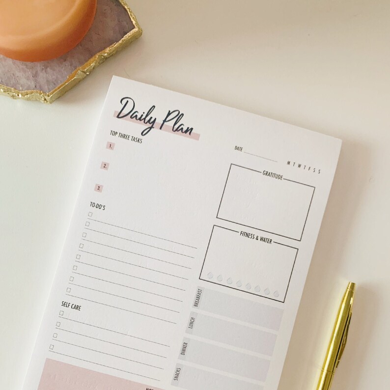K&ouml;nnte beinhalten: Ein wei&szlig;er Tagesplaner mit einem rosa Streifen und dem Text "Daily Plan" in Schwarz. Der Planer hat Abschnitte f&uuml;r die drei wichtigsten Aufgaben, To-Dos, Selbstpflege, Dankbarkeit, Fitness & Wasser, Snacks, Abendessen, Mittagessen und Fr&uuml;hst&uuml;ck.