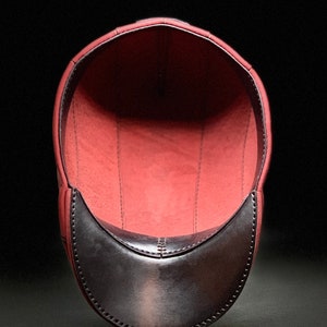 May include: A burgundy leather cap with a black leather brim. The inside of the cap is lined with a soft, red fabric.