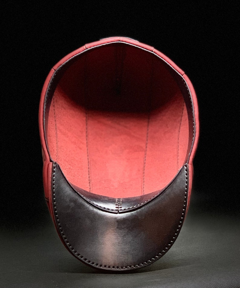 May include: A burgundy leather cap with a black leather brim. The inside of the cap is lined with a soft, red fabric.