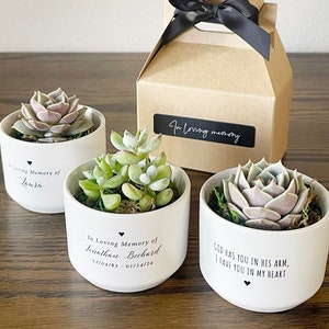 May include: Three small white ceramic pots with succulents planted inside. Each pot has a black heart and a personalized message. The first pot says "In Loving Memory of Laura". The second pot says "In Loving Memory of Jonathan Berhard 12/04/83 - 01/14/24". The third pot says "God has you in his arm. I have you in my heart."