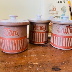 May include: Three ceramic jars with lids, each with a different label: "Jam", "Honey", and "Chutney". The jars are brown with white lids and red and white stripes.