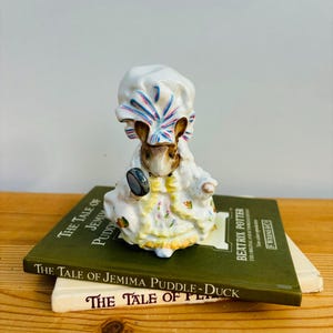 Può includere: Una statuetta in porcellana di Jemima Puddle-Duck della serie di libri per bambini di Beatrix Potter. La statuetta indossa un berretto a righe bianche e blu e un vestito giallo con dettagli floreali. La statuetta tiene uno specchio rotondo piccolo nella sua zampa destra.