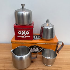 Op de afbeelding: Een set van vier keukendozen van roestvrij staal. De dozen zijn van verschillende groottes en vormen. De dozen staan op een gele en rode blikken doos met de tekst "OXO" en "The Original Beefy Cube" erop gedrukt.