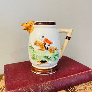 May include: A white ceramic creamer with a brown handle and a fox head spout. The creamer features a painted scene of a rider on horseback with a dog running alongside. The creamer is sitting on a red book.