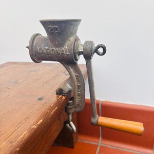 May include: An antique cast iron meat grinder, marked "NATIONAL" and "MADE IN ENGLAND", is clamped to a wooden surface. The grinder features a hand crank with a wooden handle and is a vintage kitchen tool.