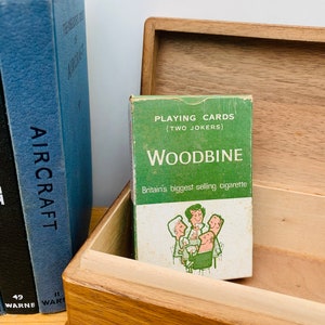 May include: A green and white box of Woodbine playing cards with two jokers. The box features a cartoon image of four people sitting around a table with drinks. The text on the box reads "Playing Cards (Two Jokers) Woodbine Britain's biggest selling cigarette."