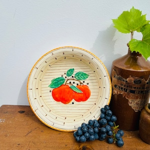 Puede incluir: Un cuenco de cerámica blanca con un borde amarillo y un diseño pintado de dos manzanas rojas con hojas verdes. El cuenco tiene un patrón de tejido de cesta y está sentado sobre una superficie de madera.