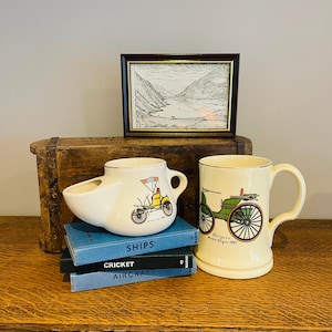 Op de afbeelding: Twee vintage keramische mokken met een geel en groen autoontwerp. De mokken staan op een stapel boeken met de titels "SHIPS", "CRICKET" en "AIRCRAFT".