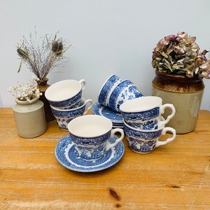 Puede incluir: Un juego de seis tazas de té y platillos azules y blancos con un patrón chino tradicional. Las tazas y los platillos están dispuestos sobre una mesa de madera.