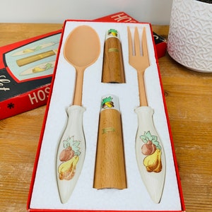 Può includere: Un set di tre utensili da cucina vintage del marchio Hostess in una scatola rossa. Il set comprende un cucchiaio, una forchetta e un piccolo contenitore di legno. Gli utensili sono in plastica e hanno un manico in ceramica bianca con un motivo a frutta. La scatola è rivestita di schiuma bianca.