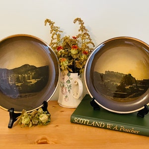 Peut inclure: Deux assiettes décoratives avec une bordure dorée représentant des photographies vintage en noir et blanc d'Édimbourg, en Écosse. Les assiettes sont exposées sur des supports noirs et un livre vert intitulé "Scotland W.A. Poucher" se trouve au premier plan.