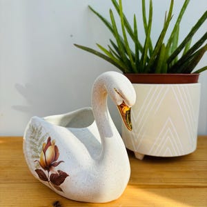 Könnte beinhalten: Weißer Keramik-Schwanen-Pflanztopf mit braunem und goldenem Blumendesign. Der Schwan hat einen goldenen Schnabel und steht auf einer Holzoberfläche.