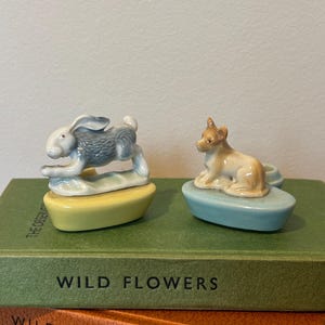 May include: Two ceramic animal figurines: a gray and white rabbit and a tan dog. The rabbit is on a yellow base, and the dog is on a blue base. Both are placed on top of green books with the titles "Wild Flowers" and "Wild Animals".
