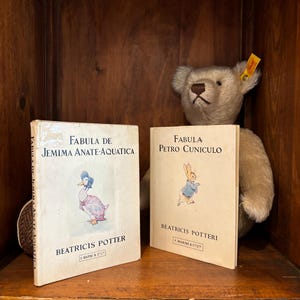 Puede incluir: Se muestran dos libros infantiles antiguos, "Fabula de Jemima Anate-Aquatica" y "Fabula Petro Cuniculo" de Beatricis Potter. Un oso de peluche está colocado detrás de los libros. Los libros tienen ilustraciones de un pato y un conejo.